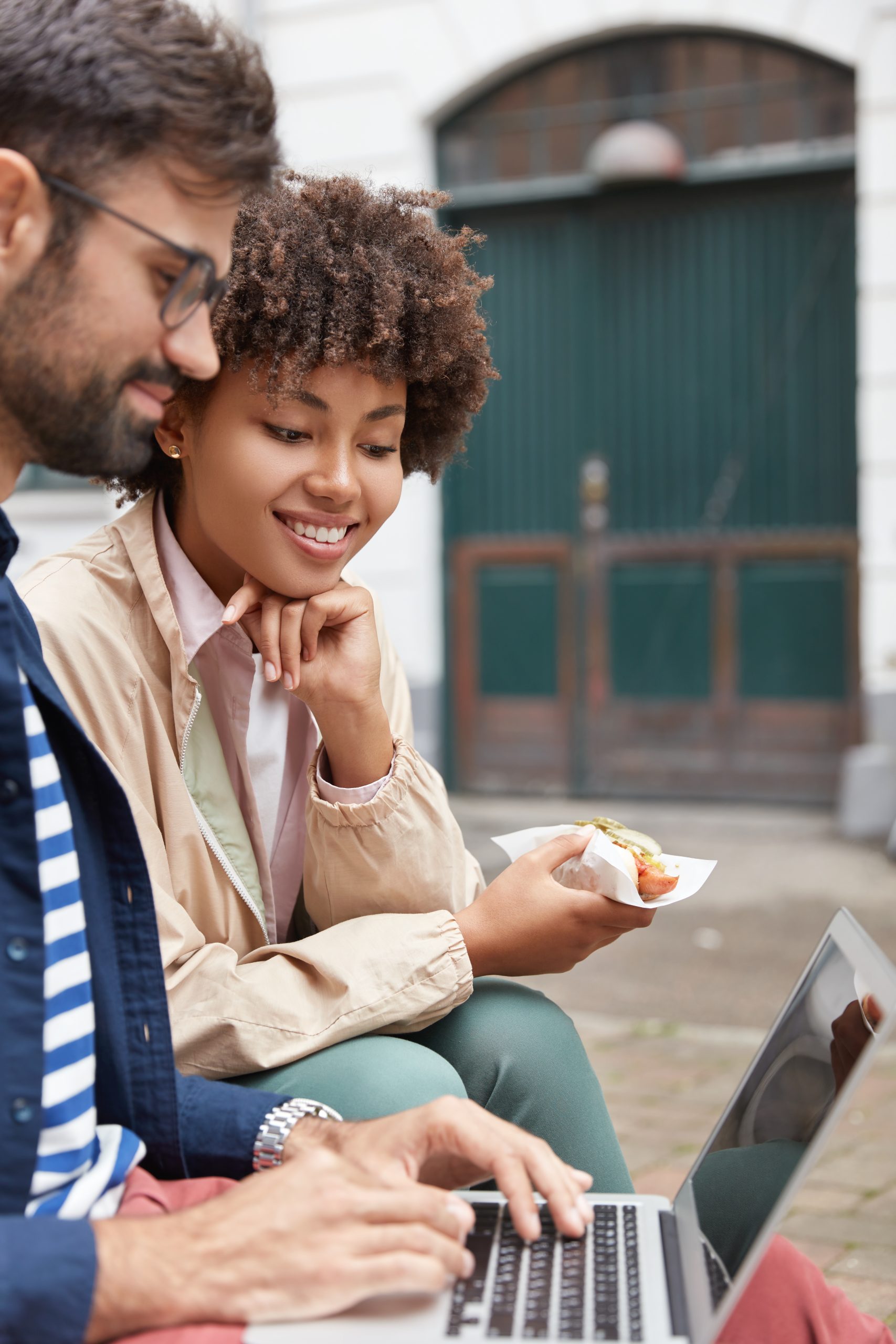 People, lifestyle and networking concept. Vertical shot of mixed race woman and man use laptop computer for e learning, keyboard text message, pose at street with sandwich, have positive expressions